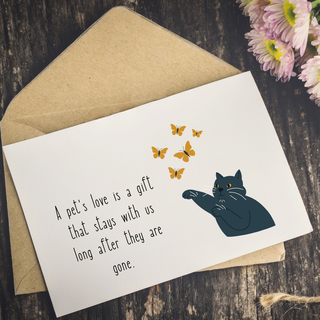 A white greeting card with a black cat illustration and yellow butterflies, placed on top of a kraft paper envelope on a dark wooden surface with pink flowers in the corner.
