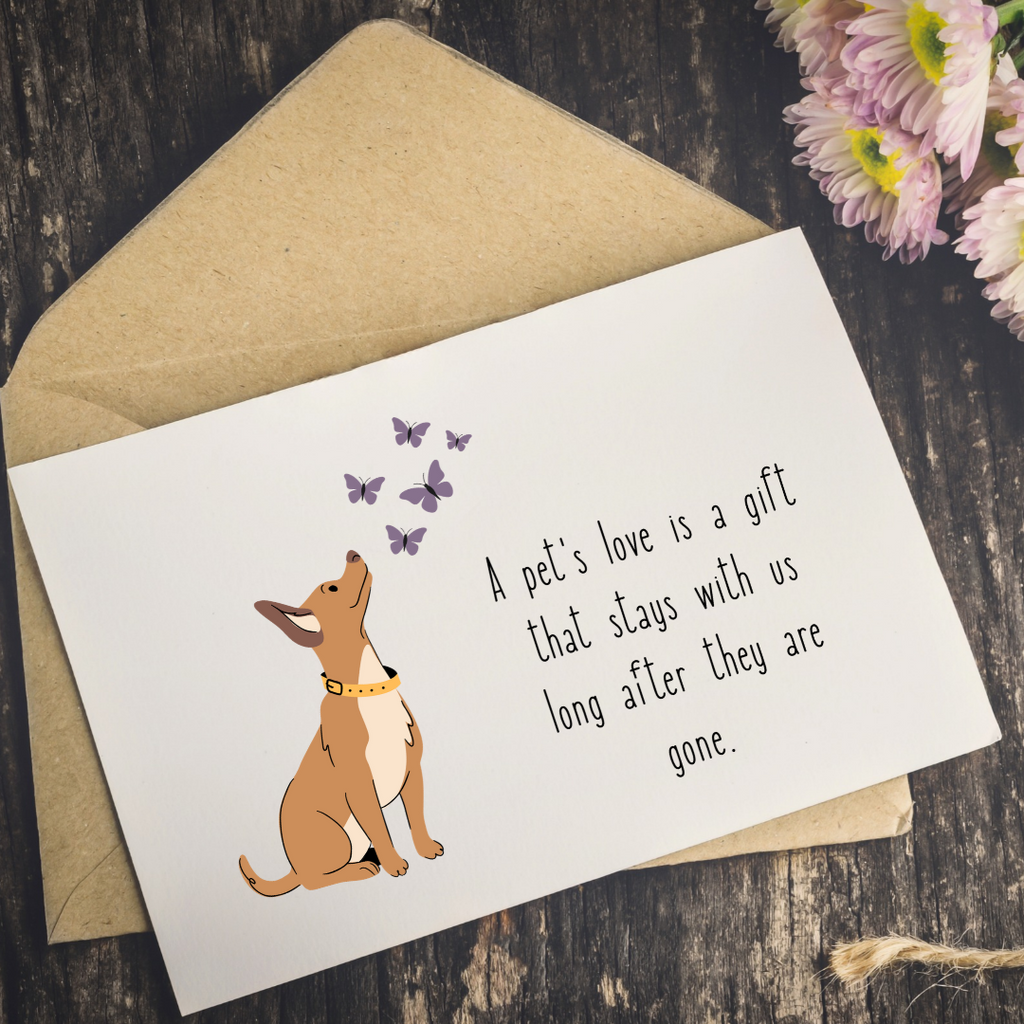 A square white greeting card with an illustration of a brown dog sitting among purple butterflies and the printed quote, 'A pet's love is a gift that stays with us long after they are gone.' The card is placed on top of a kraft paper envelope on a dark wood surface, with pink and white flowers in the corner.
