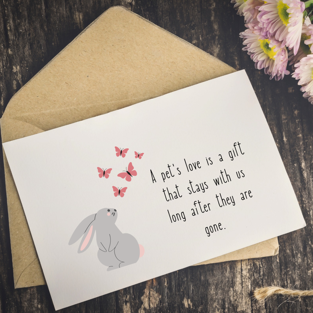 A white greeting card with a gray bunny illustration, pink butterflies, and the text 'A pet's love is a gift that stays with us long after they are gone,' placed on top of a kraft paper envelope on a dark wood surface with flowers in the corner.