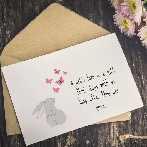 A white greeting card with a gray bunny illustration, pink butterflies, and the text 'A pet's love is a gift that stays with us long after they are gone,' placed on top of a kraft paper envelope on a dark wood surface with flowers in the corner.