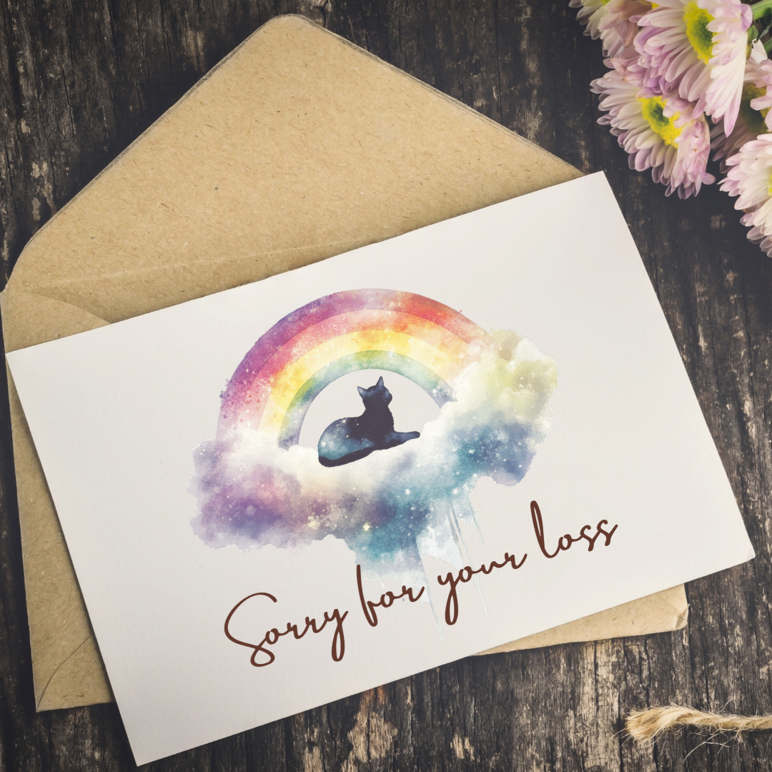 A white memorial card with a watercolor rainbow, cat silhouette, and the words 'Sorry for your loss' in brown script, placed on top of a kraft envelope on a dark wood surface with flowers in the corner.