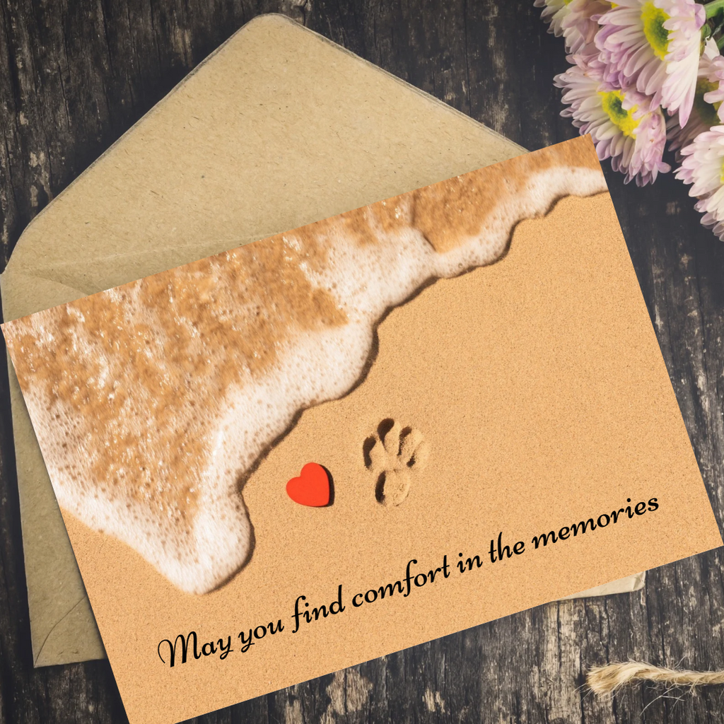 A square memorial card with a kraft-paper background and a printed beach scene, shown with a matching kraft envelope on a dark wood surface. The card features a foam shoreline, a small red heart, a paw print, and the text 'May you find comfort in the memories.