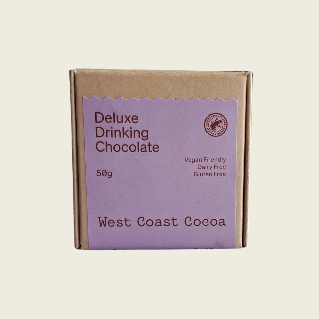 A square kraft paper box of West Coast Cocoa Deluxe Drinking Chocolate, 50g, featuring a lavender purple label with brown text, set against a solid cream background.