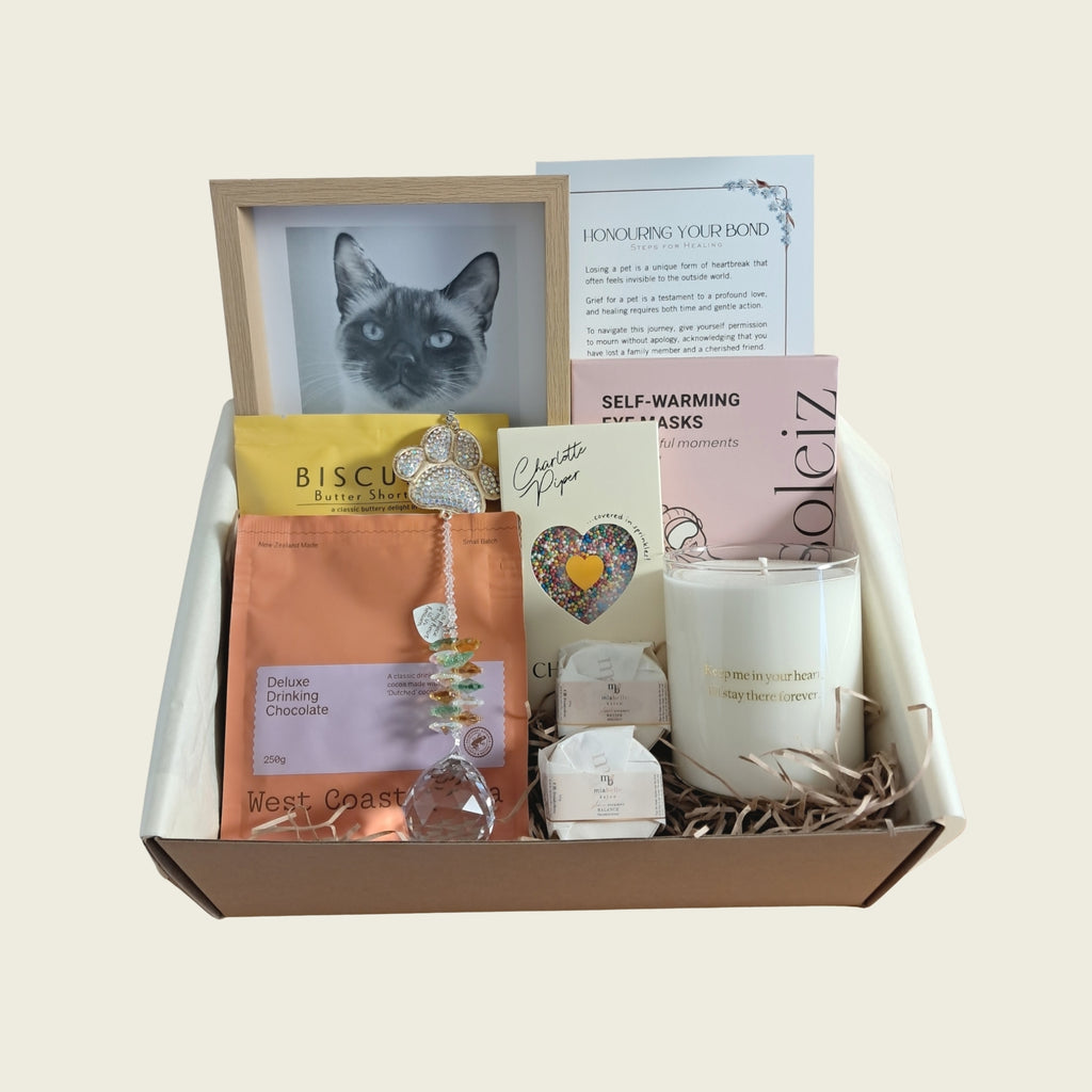 A curated gift box set containing a framed cat portrait, a candle, chocolate, shower steamers, a crystal bead charm, and printed cards, arranged in a brown box with shredded paper on a light beige background.