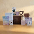 A curated gift set arranged on a plain light beige background. The set includes a brown box labeled Rainbow Bridge Comfort and 'thinking of you,' a candle, a pink box, a card, a small sachet, a heart-shaped wooden ornament, and several printed cards with memorial or condolence text.