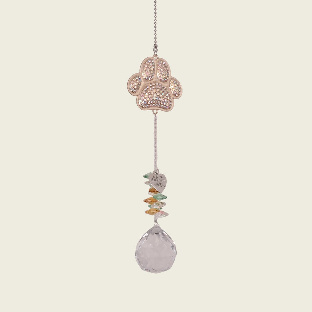 A hanging suncatcher ornament featuring a gold paw print filled with small clear rhinestones, a string of colorful flat stones, and a faceted clear teardrop crystal at the bottom, shown against a solid light beige background.