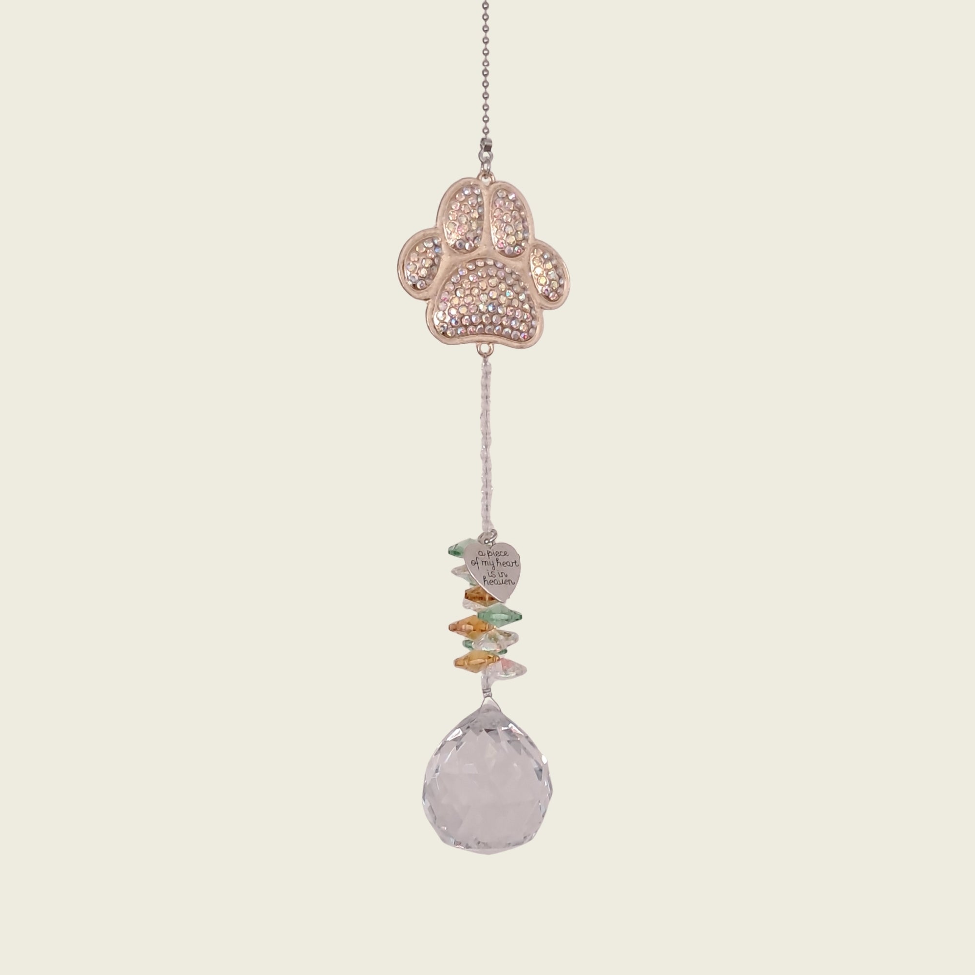 A hanging suncatcher ornament featuring a gold paw print filled with small clear rhinestones, a string of colorful flat stones, and a faceted clear teardrop crystal at the bottom, shown against a solid light beige background.