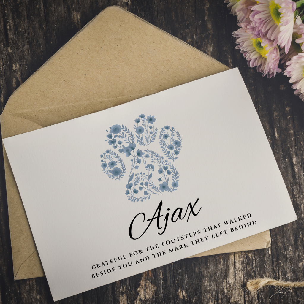 A white memorial or sympathy card with a blue floral illustration and the name 'Ajax' in black script, placed on top of a kraft paper envelope on a dark wood surface with pink flowers in the corner.