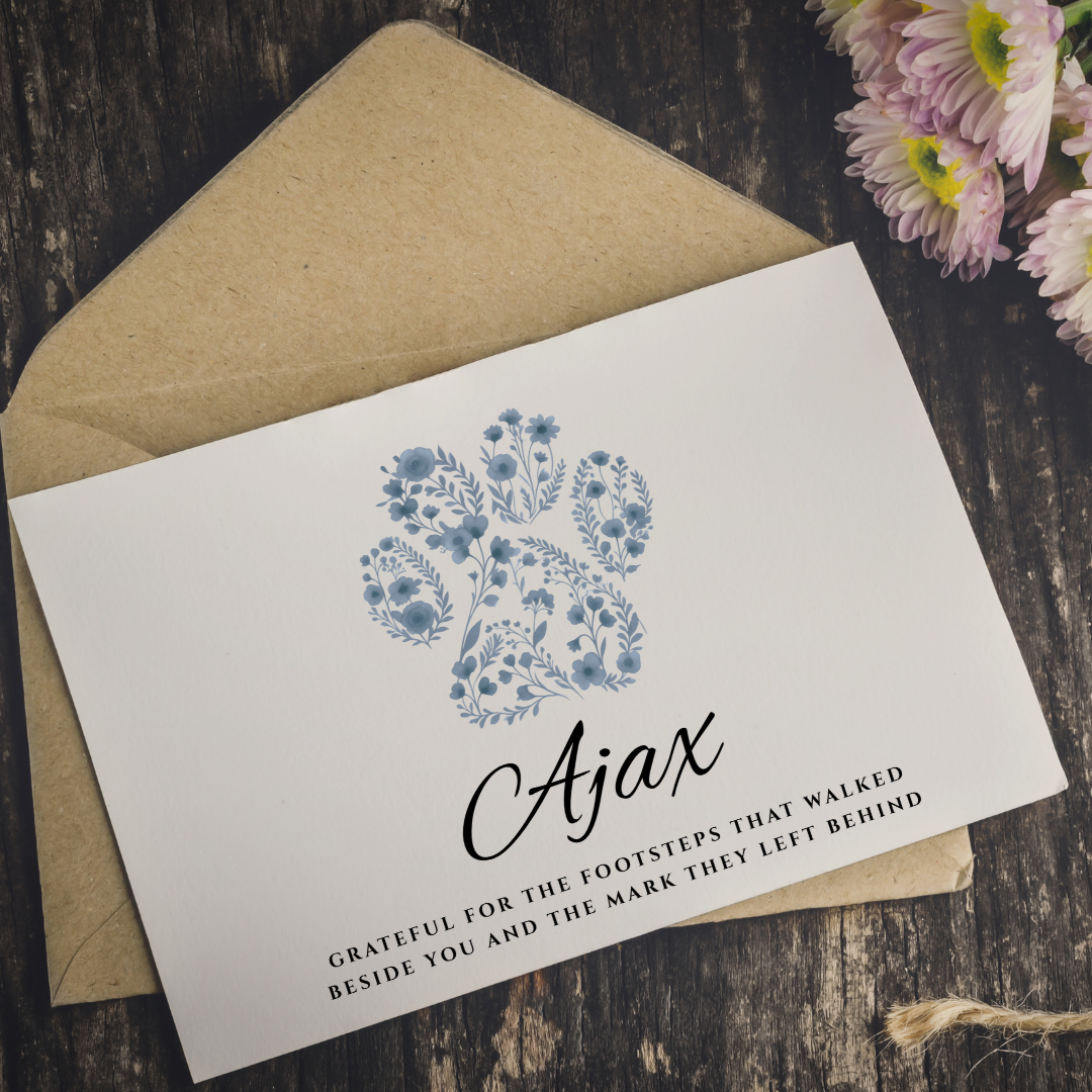 A white memorial or sympathy card with a blue floral illustration and the name 'Ajax' in black script, placed on top of a kraft paper envelope on a dark wood surface with pink flowers in the corner.