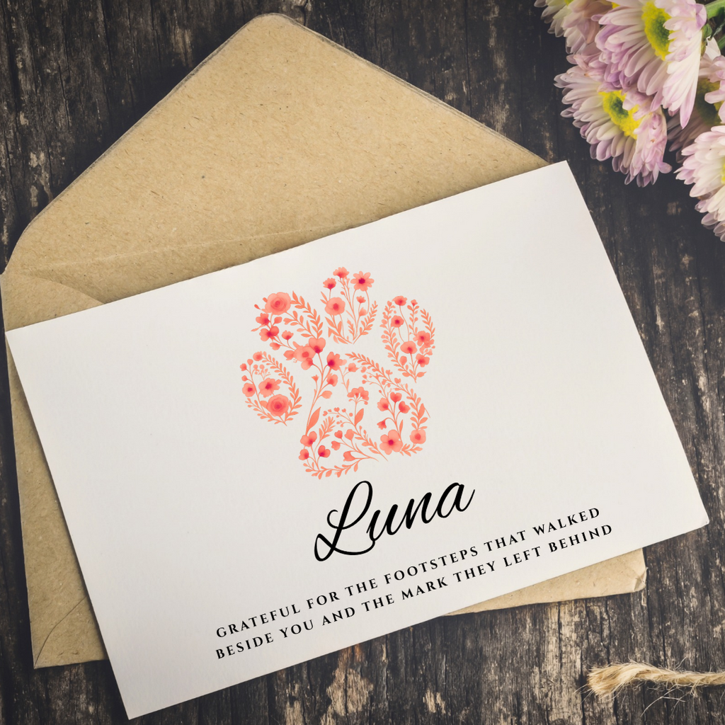 A white printed card with a coral floral illustration and the name 'Luna' in black script, placed on top of a kraft paper envelope on a dark wood surface with pink flowers in the corner.