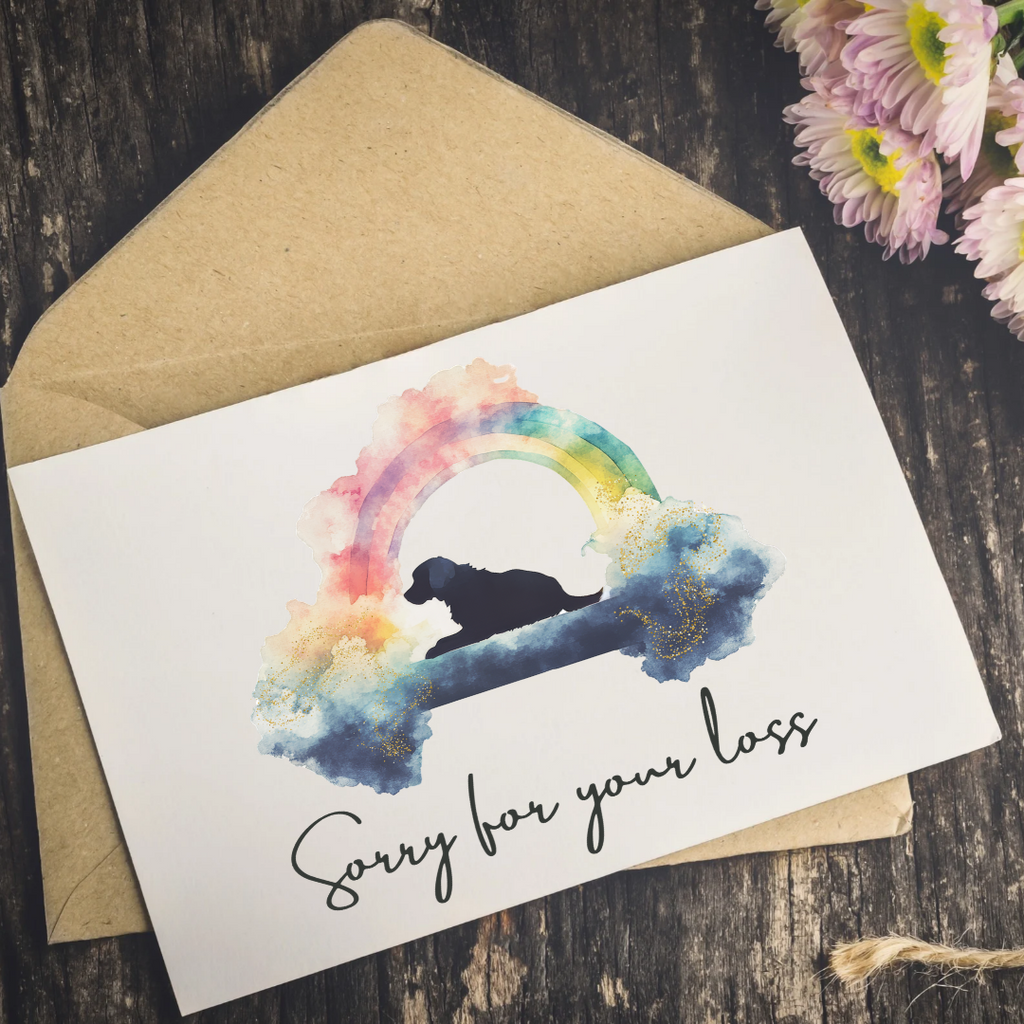 A white memorial card with a watercolor rainbow and dog silhouette, placed on top of a kraft envelope on a dark wood surface with pink flowers at the corner.