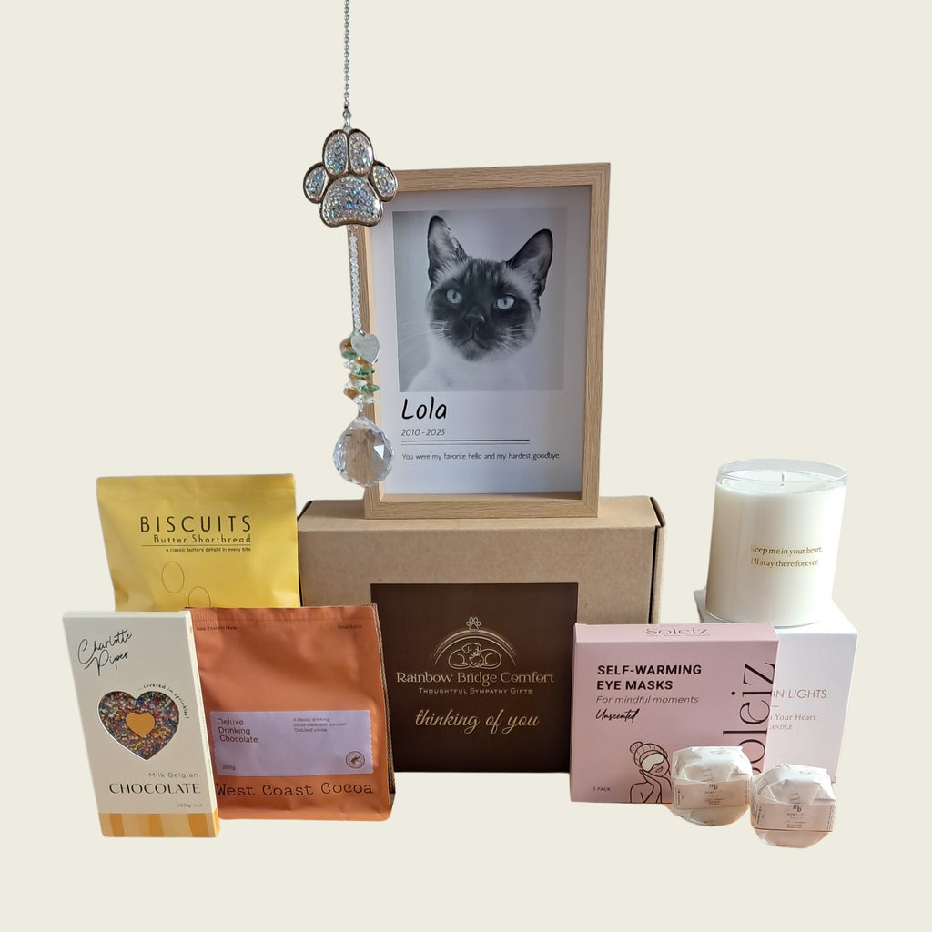 A curated gift basket set featuring a custom cat portrait in a wooden frame, a silver key charm, a white candle, self-warming eye masks, chocolate and coffee pouches, and shower steamers, arranged on a light beige background.