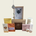A curated gift basket set featuring a custom cat portrait in a wooden frame, a silver key charm, a white candle, self-warming eye masks, chocolate and coffee pouches, and shower steamers, arranged on a light beige background.