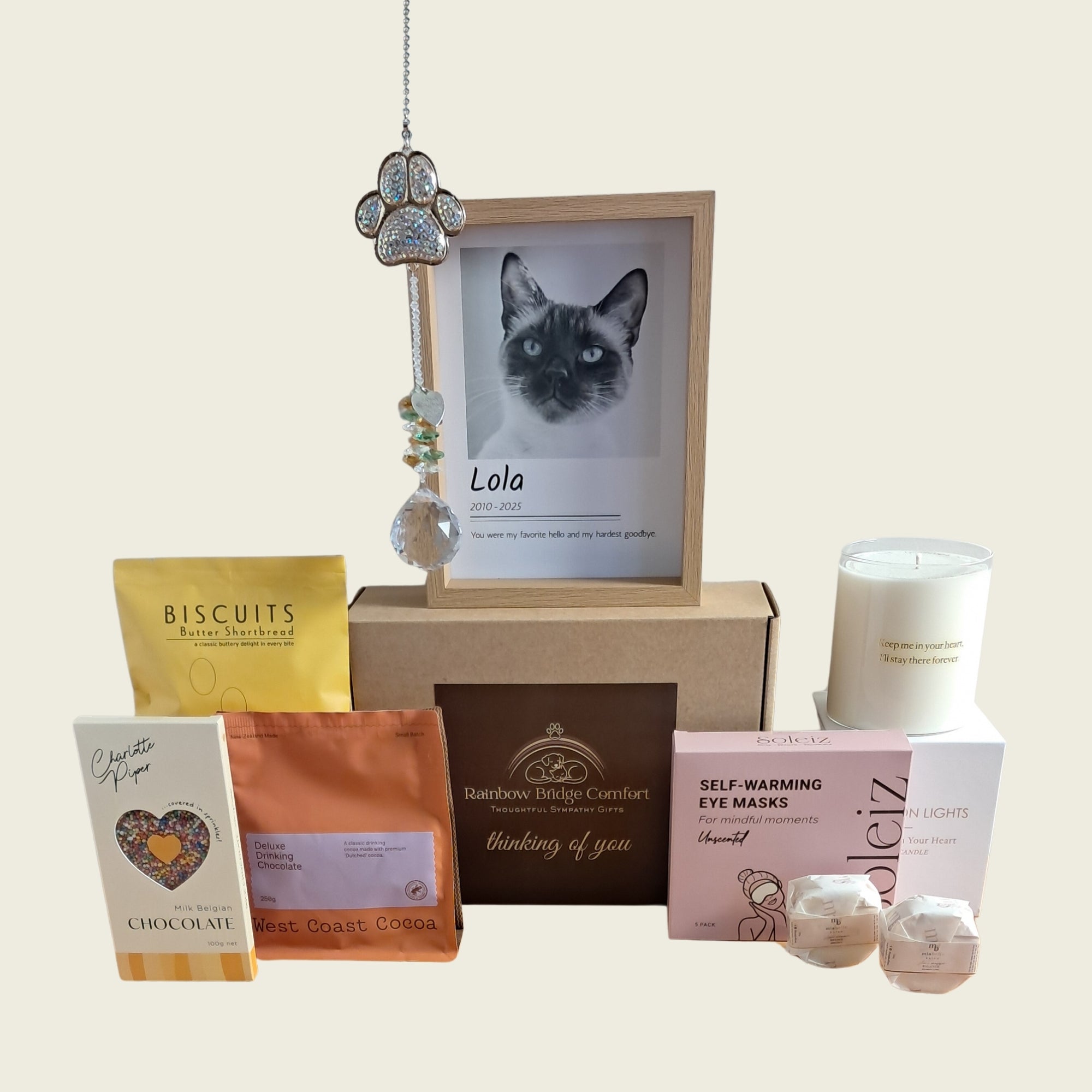 A curated gift basket set featuring a custom cat portrait in a wooden frame, a silver key charm, a white candle, self-warming eye masks, chocolate and coffee pouches, and shower steamers, arranged on a light beige background.
