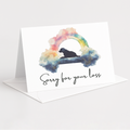 A white greeting card standing on its front flap, with a black script phrase reading 'Sorry for your loss' and a watercolor-style rainbow arch over a black dog silhouette. A white envelope is placed behind the card on a light gray background.