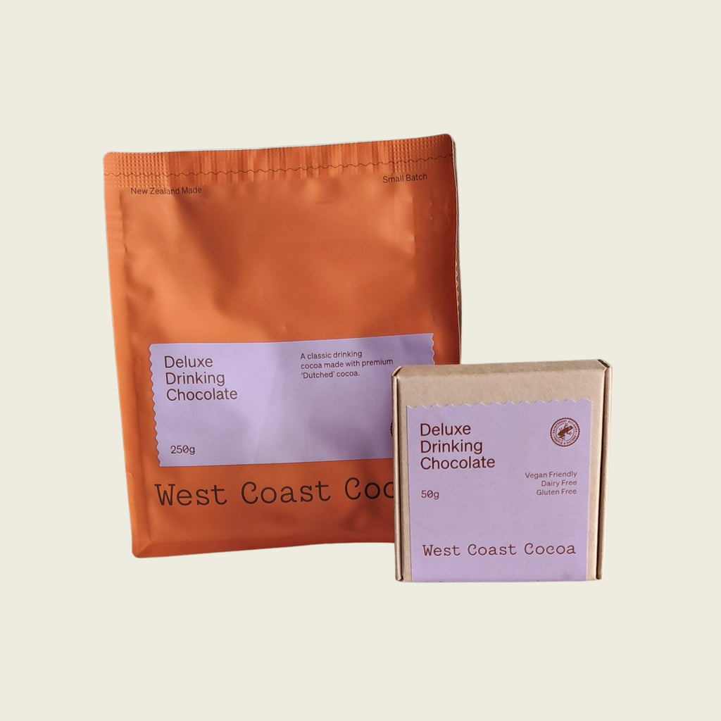 Two West Coast Cocoa packages on a light beige background: a large orange foil bag labeled 'Deluxe Drinking Chocolate 250g' and a smaller kraft paper box labeled 'Deluxe Drinking Chocolate 150g'.
