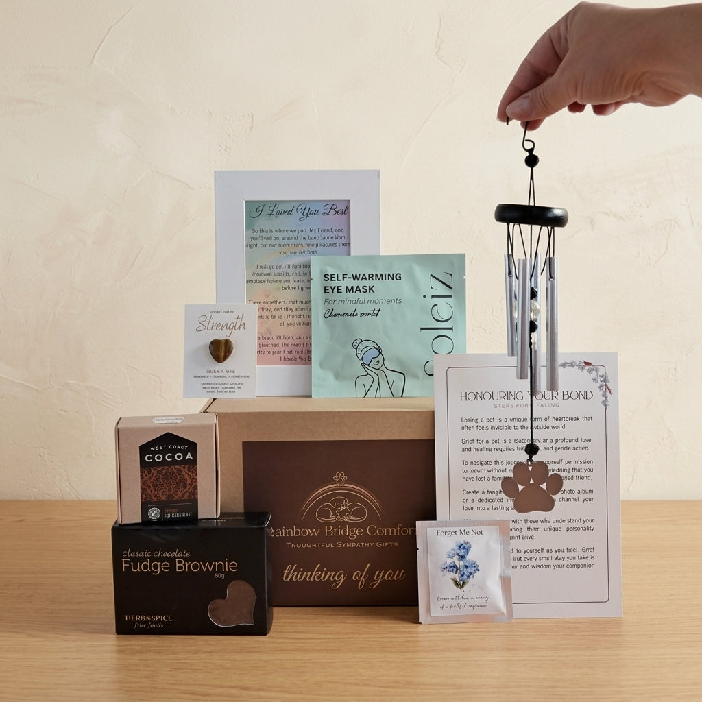 Rainbow Bridge Comfort Essential Comforts Pet Loss Sympathy Gift Box featuring a paw-print wind chime, gourmet treats, Tiger's Eye pocket heart, and memorial keepsakes in a branded kraft gift box.