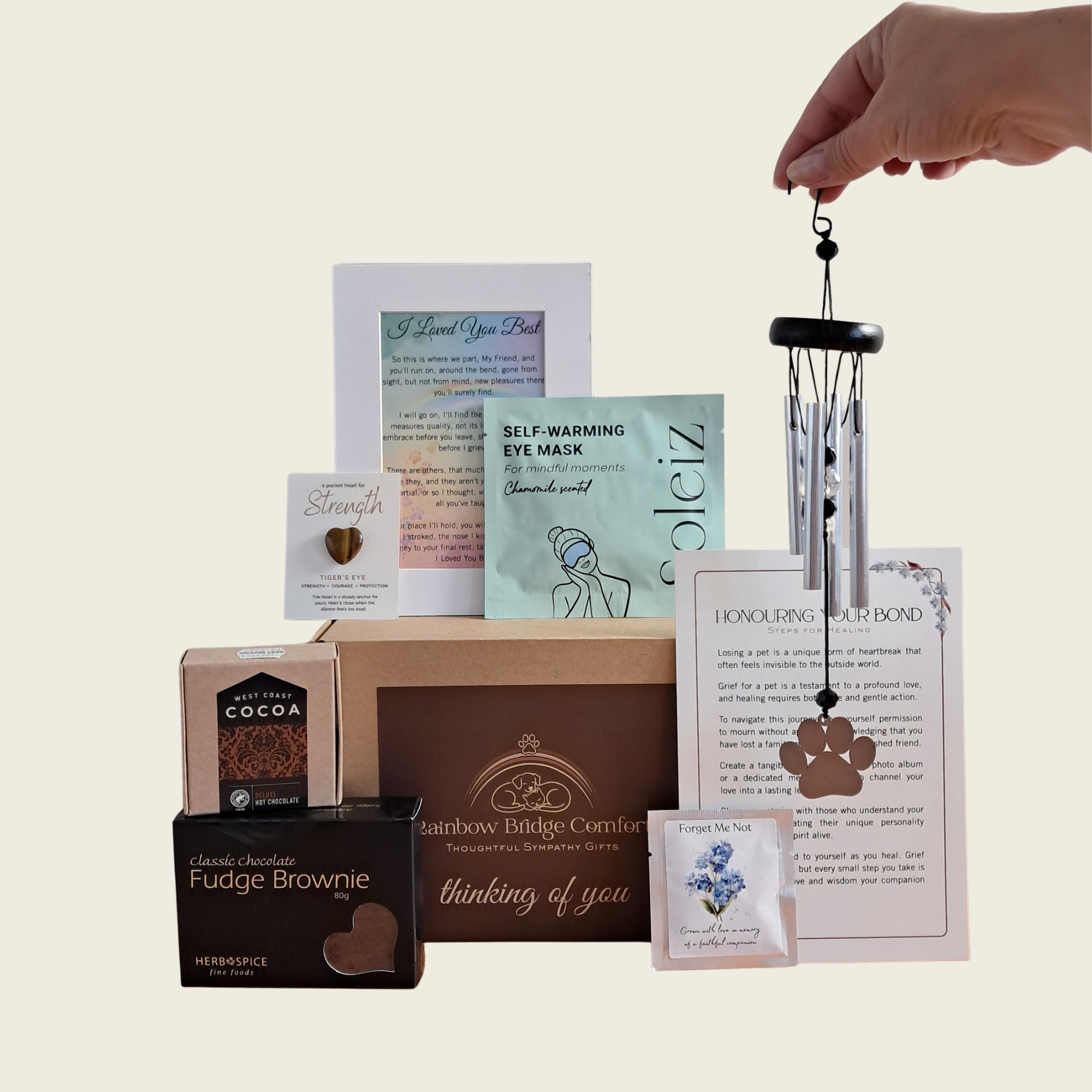 Rainbow Bridge Comfort Essential Comforts Pet Loss Sympathy Gift Box featuring a paw-print wind chime, gourmet treats, Tiger's Eye pocket heart, and memorial keepsakes in a branded kraft gift box.