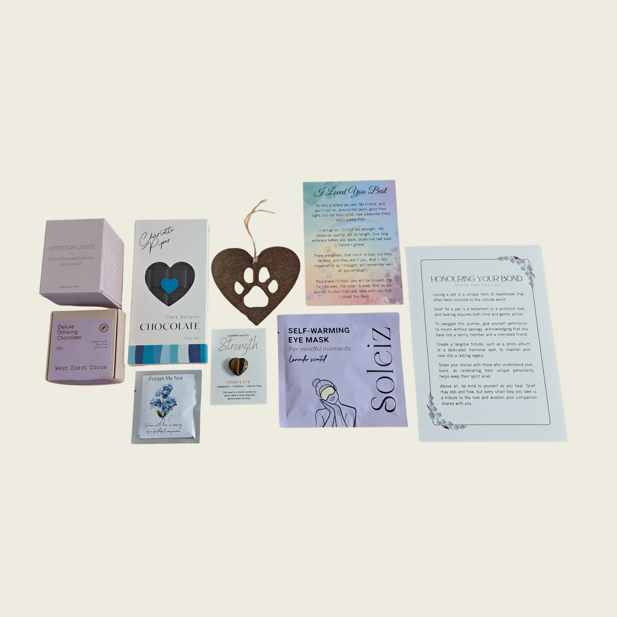 A flat-lay arrangement of multiple small product items and printed cards on a light warm-gray background. Visible items include a pink box, a chocolate bar package, a heart-shaped tag, a self-warming eye mask packet, a sachet, and two printed cards with text.
