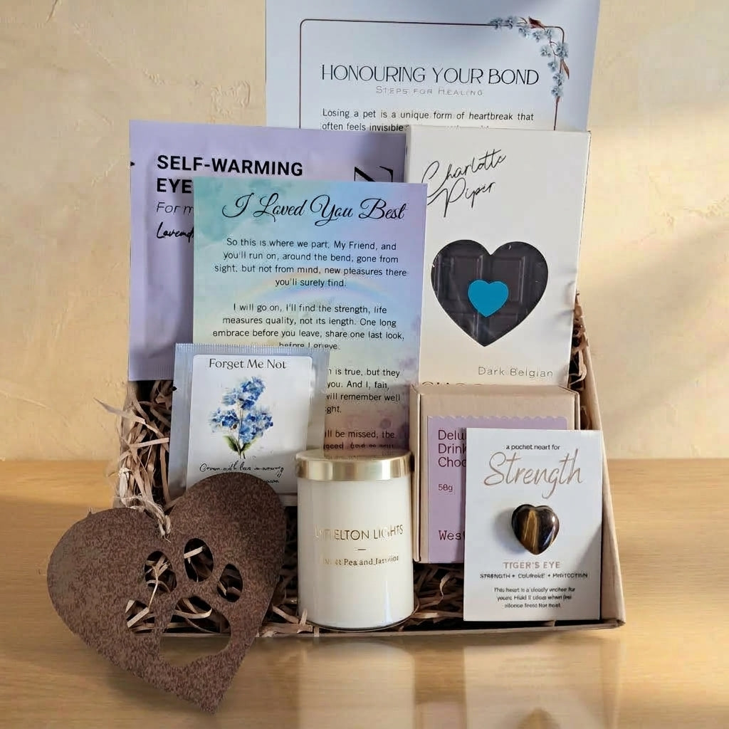 A memorial gift basket arrangement containing a white candle, printed cards with messages, a dark blue heart-shaped keepsake, a wooden heart-shaped ornament, and a printed booklet, all arranged in a basket with shredded paper filler against a light beige background.