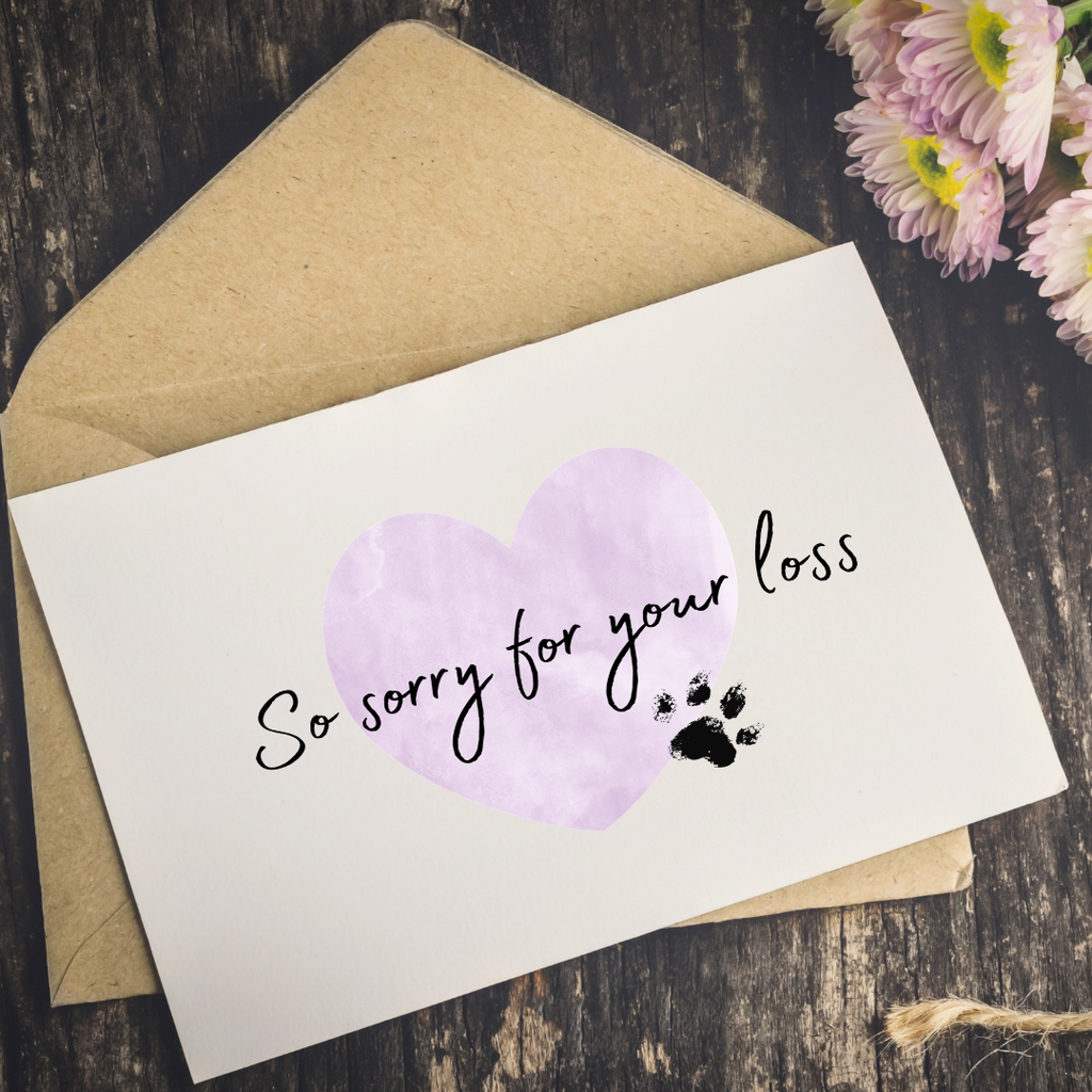A white memorial card with a watercolor-style lavender heart, black script text reading 'So sorry for your loss,' and a small black paw print, placed on top of a kraft paper envelope on a dark wood surface with pink flowers in the corner.