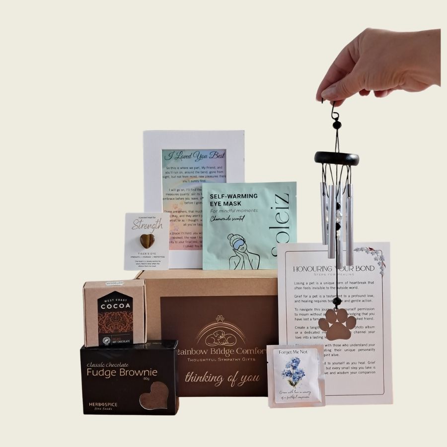 Rainbow Bridge Comfort Essential Comforts Pet Loss Sympathy Gift Box featuring a paw-print wind chime, gourmet treats, Tiger's Eye pocket heart, and memorial keepsakes in a branded kraft gift box.